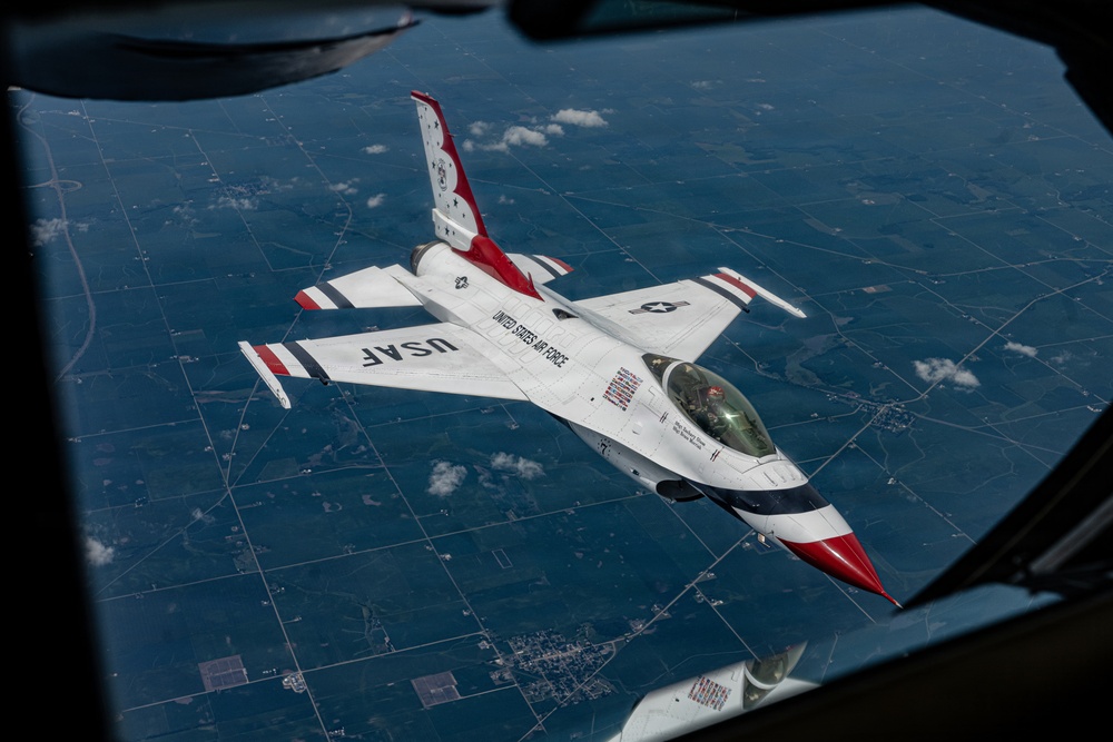 An aerial photograph taken from inside a larger aircraft showing a U.S. Air Force Thunderbirds F-16 Fighting Falcon in close proximity, likely during an air-to-air refueling or formation flight. The aircraft displays the distinctive Thunderbirds livery of white with red and blue accents, with 'USAF' and 'United States Air Force' markings clearly visible. A patchwork of agricultural fields is visible far below, indicating high-altitude flight.