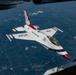 An aerial photograph taken from inside a larger aircraft showing a U.S. Air Force Thunderbirds F-16 Fighting Falcon in close proximity, likely during an air-to-air refueling or formation flight. The aircraft displays the distinctive Thunderbirds livery of white with red and blue accents, with 'USAF' and 'United States Air Force' markings clearly visible. A patchwork of agricultural fields is visible far below, indicating high-altitude flight.