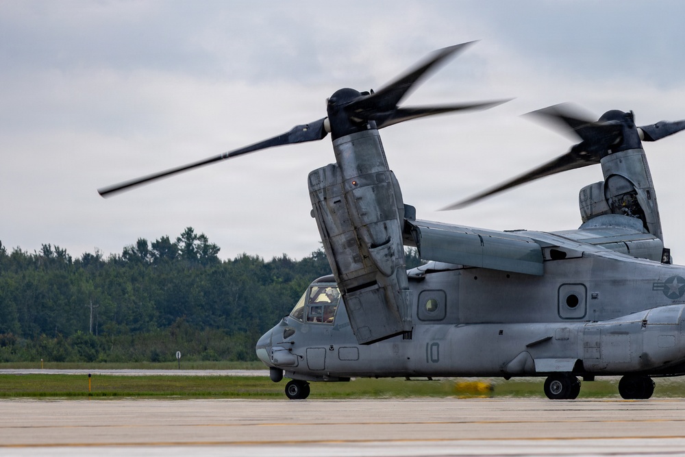 A U.S. military V-22 Osprey tiltrotor aircraft is shown on a tarmac with its rotors spinning, indicating it is either preparing for takeoff or has recently landed. The aircraft bears the number '10' on its fuselage and displays U.S. military insignia. The setting appears to be a military airfield surrounded by tree lines under an overcast sky.