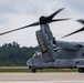 A U.S. military V-22 Osprey tiltrotor aircraft is shown on a tarmac with its rotors spinning, indicating it is either preparing for takeoff or has recently landed. The aircraft bears the number '10' on its fuselage and displays U.S. military insignia. The setting appears to be a military airfield surrounded by tree lines under an overcast sky.