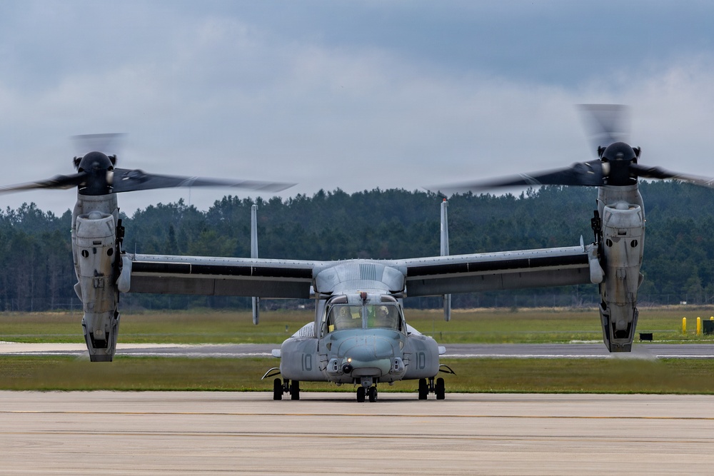A military tiltrotor aircraft, identified as a Bell Boeing V-22 Osprey, is taxiing on a runway with both rotors spinning in helicopter mode. The aircraft bears the number '10' on its nose and appears to be in U.S. Marine Corps livery. The setting is a military airfield with a treeline and overcast sky in the background.