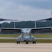 A military tiltrotor aircraft, identified as a Bell Boeing V-22 Osprey, is taxiing on a runway with both rotors spinning in helicopter mode. The aircraft bears the number '10' on its nose and appears to be in U.S. Marine Corps livery. The setting is a military airfield with a treeline and overcast sky in the background.