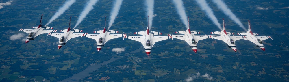 A formation of USAF Thunderbirds aerobatic demonstration jets fly in a tight echelon formation high above a patchwork landscape. All aircraft are emitting white smoke trails from their engines, indicating an active aerial demonstration. The jets are identifiable as F-16 Fighting Falcons painted in the iconic red, white, and blue Thunderbirds livery.