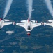 A formation of USAF Thunderbirds aerobatic demonstration jets fly in a tight echelon formation high above a patchwork landscape. All aircraft are emitting white smoke trails from their engines, indicating an active aerial demonstration. The jets are identifiable as F-16 Fighting Falcons painted in the iconic red, white, and blue Thunderbirds livery.