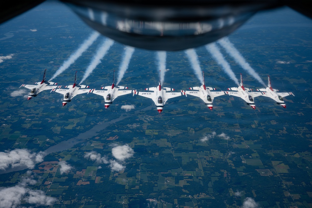 Seven USAF Thunderbirds aerobatic demonstration jets fly in a tight echelon formation, photographed from above through the belly window of a larger aircraft, likely a KC-135 tanker. Each F-16 Fighting Falcon displays the distinctive red, white, and blue Thunderbirds livery with 'USAF' markings and produces white contrails. The formation flies over a patchwork of agricultural fields and scattered clouds at high altitude.