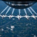 Seven USAF Thunderbirds aerobatic demonstration jets fly in a tight echelon formation, photographed from above through the belly window of a larger aircraft, likely a KC-135 tanker. Each F-16 Fighting Falcon displays the distinctive red, white, and blue Thunderbirds livery with 'USAF' markings and produces white contrails. The formation flies over a patchwork of agricultural fields and scattered clouds at high altitude.
