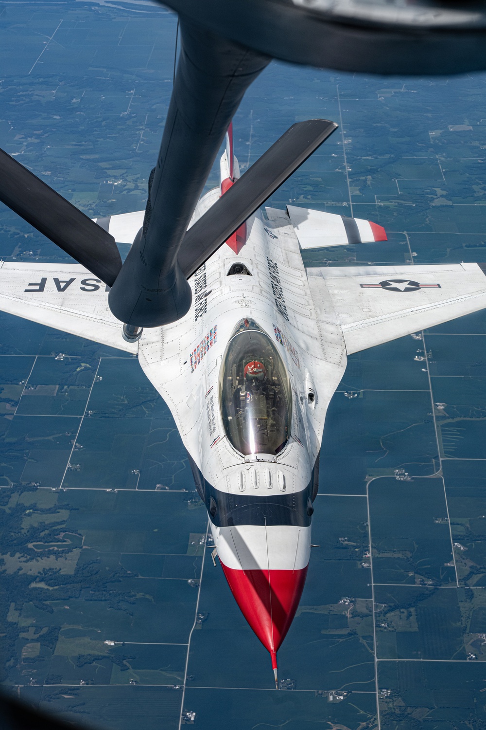 An aerial photograph taken from a tanker aircraft showing a USAF Thunderbirds F-16 Fighting Falcon during an aerial refueling operation. The fighter jet is viewed from directly above and behind, with the tanker's refueling boom extending toward the aircraft's receptacle. The patchwork agricultural landscape below confirms the aircraft are flying at altitude over a rural area.