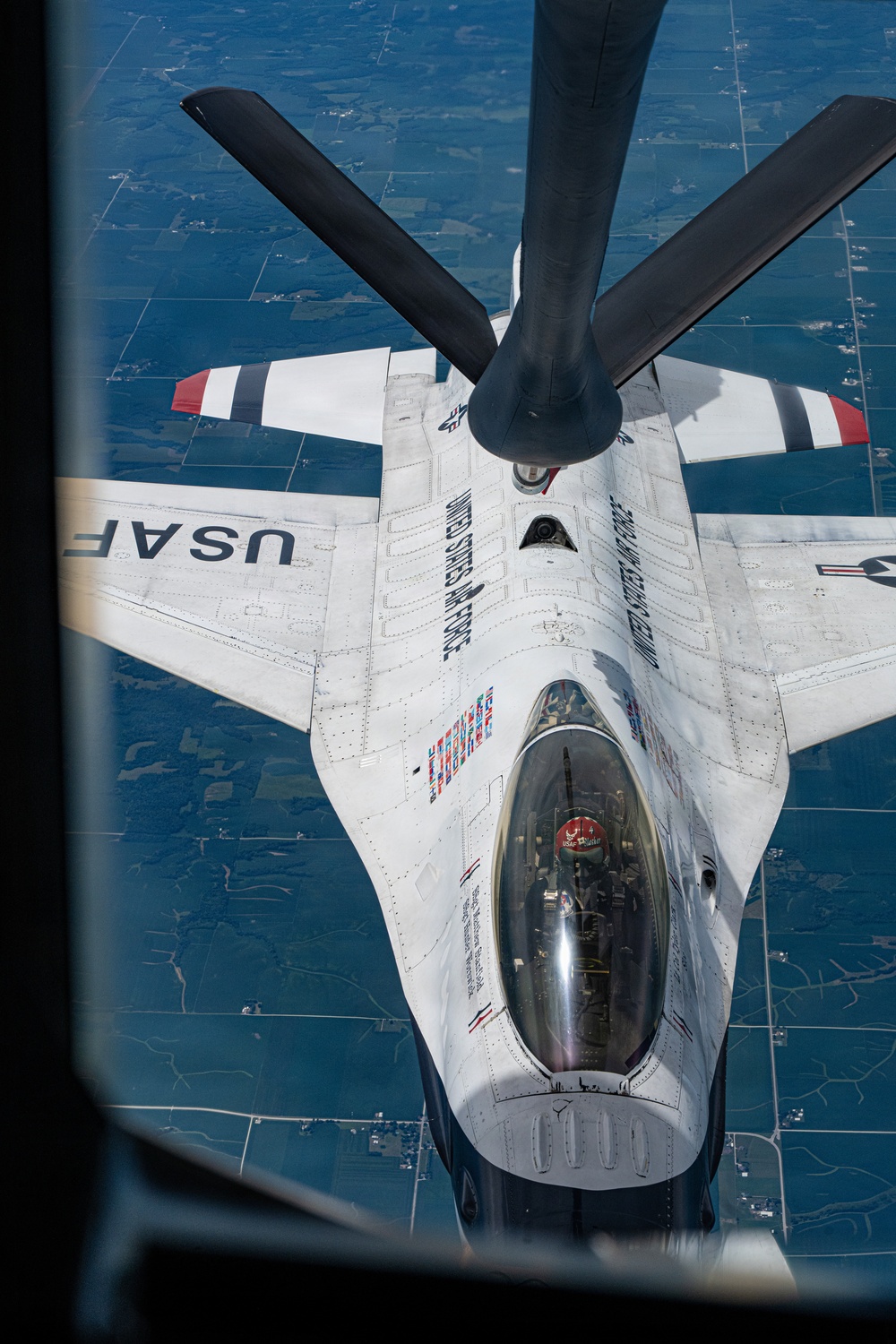 An aerial refueling operation is captured from the boom operator's perspective aboard a tanker aircraft, showing a USAF Thunderbirds F-16 Fighting Falcon receiving fuel mid-flight. The fighter jet is painted in the distinctive white, red, and blue Thunderbirds demonstration team livery with 'UNITED STATES AIR FORCE' markings. The ground below shows a flat, agricultural landscape with a grid pattern of fields visible thousands of feet below.