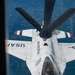 An aerial refueling operation is captured from the boom operator's perspective aboard a tanker aircraft, showing a USAF Thunderbirds F-16 Fighting Falcon receiving fuel mid-flight. The fighter jet is painted in the distinctive white, red, and blue Thunderbirds demonstration team livery with 'UNITED STATES AIR FORCE' markings. The ground below shows a flat, agricultural landscape with a grid pattern of fields visible thousands of feet below.