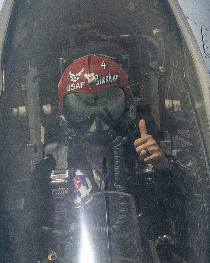 A USAF pilot is seated in a military jet cockpit, fully suited in flight gear including an oxygen mask and a red helmet. The helmet is marked with 'USAF', the Air Force wings insignia, the call sign 'Slasher', and the number '4'. The pilot is giving a thumbs-up gesture, visible through the closed canopy.