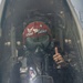 A USAF pilot is seated in a military jet cockpit, fully suited in flight gear including an oxygen mask and a red helmet. The helmet is marked with 'USAF', the Air Force wings insignia, the call sign 'Slasher', and the number '4'. The pilot is giving a thumbs-up gesture, visible through the closed canopy.