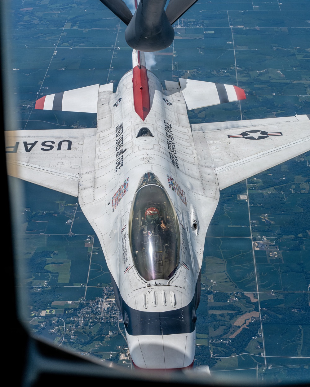 An aerial photograph taken from a tanker aircraft showing a U.S. Air Force Thunderbirds F-16 Fighting Falcon during an aerial refueling operation. The jet is viewed from directly above and behind, with the refueling boom visible at the top of the frame. The pilot is visible through the bubble canopy and appears to be giving a thumbs-up gesture.