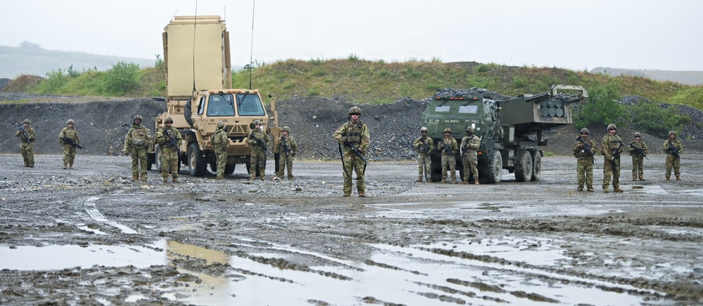 A group of approximately 15 military personnel in combat uniforms are standing in formation on a muddy terrain during overcast/rainy conditions. They are positioned in front of two military vehicles, including what appears to be a radar system mounted on a tactical truck and a multiple launch rocket system (MLRS). The scene appears to be a military exercise, demonstration, or unit photo opportunity at a training area.