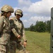 Two female military personnel in camouflage uniforms are standing near a shooting range structure, appearing to discuss or review something. One soldier wears a combat helmet while the other wears hearing protection (earmuffs), suggesting they are at or near an active firing range. A tower structure and tree line are visible in the background.