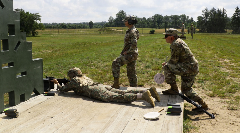 Military personnel are conducting a rifle marksmanship training exercise at an outdoor firing range. One soldier is in the prone position actively firing downrange, while two others stand and crouch nearby in an observer or safety officer role. Additional personnel are visible in the background at other firing positions.