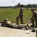 Military personnel are conducting a rifle marksmanship training exercise at an outdoor firing range. One soldier is in the prone position actively firing downrange, while two others stand and crouch nearby in an observer or safety officer role. Additional personnel are visible in the background at other firing positions.
