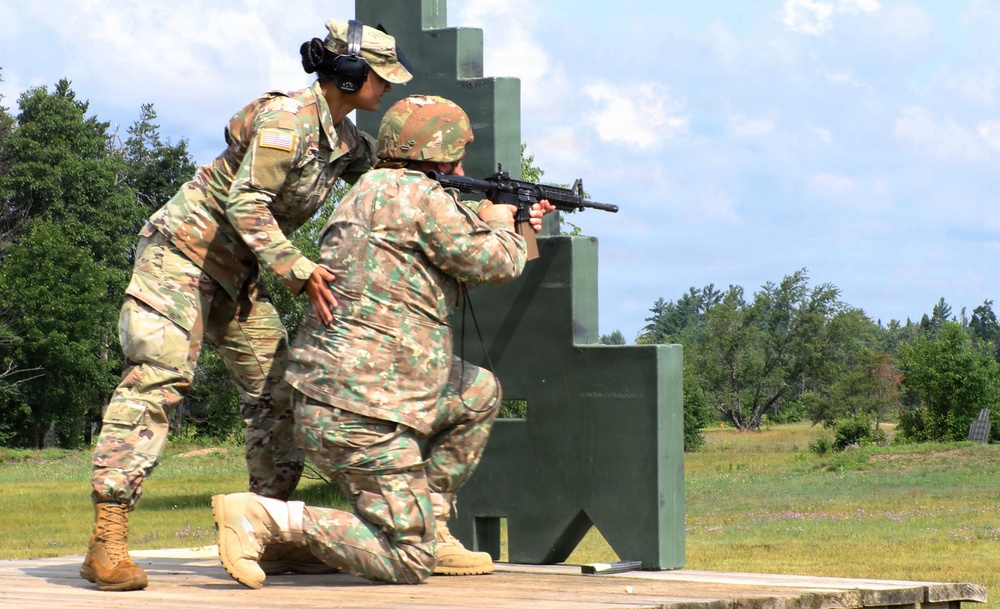 Two military personnel are engaged in a weapons training exercise at an outdoor firing range. One soldier in U.S. Army OCP (Operational Camouflage Pattern) uniform is providing instruction or coaching to a second soldier who is in a kneeling firing position aiming an M4/M16-style rifle. The setting appears to be a standard military qualification range with a green shooting barricade structure.