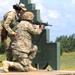 Two military personnel are engaged in a weapons training exercise at an outdoor firing range. One soldier in U.S. Army OCP (Operational Camouflage Pattern) uniform is providing instruction or coaching to a second soldier who is in a kneeling firing position aiming an M4/M16-style rifle. The setting appears to be a standard military qualification range with a green shooting barricade structure.