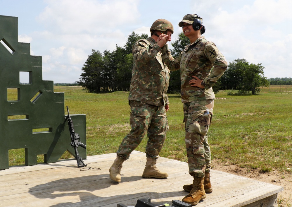Two military personnel in camouflage uniforms are standing on a wooden platform at what appears to be a firing range. One soldier wearing a helmet is gesturing toward the face of the other soldier who is wearing a cap and hearing protection. A rifle is leaned against a green target structure to their left.