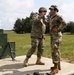 Two military personnel in camouflage uniforms are standing on a wooden platform at what appears to be a firing range. One soldier wearing a helmet is gesturing toward the face of the other soldier who is wearing a cap and hearing protection. A rifle is leaned against a green target structure to their left.