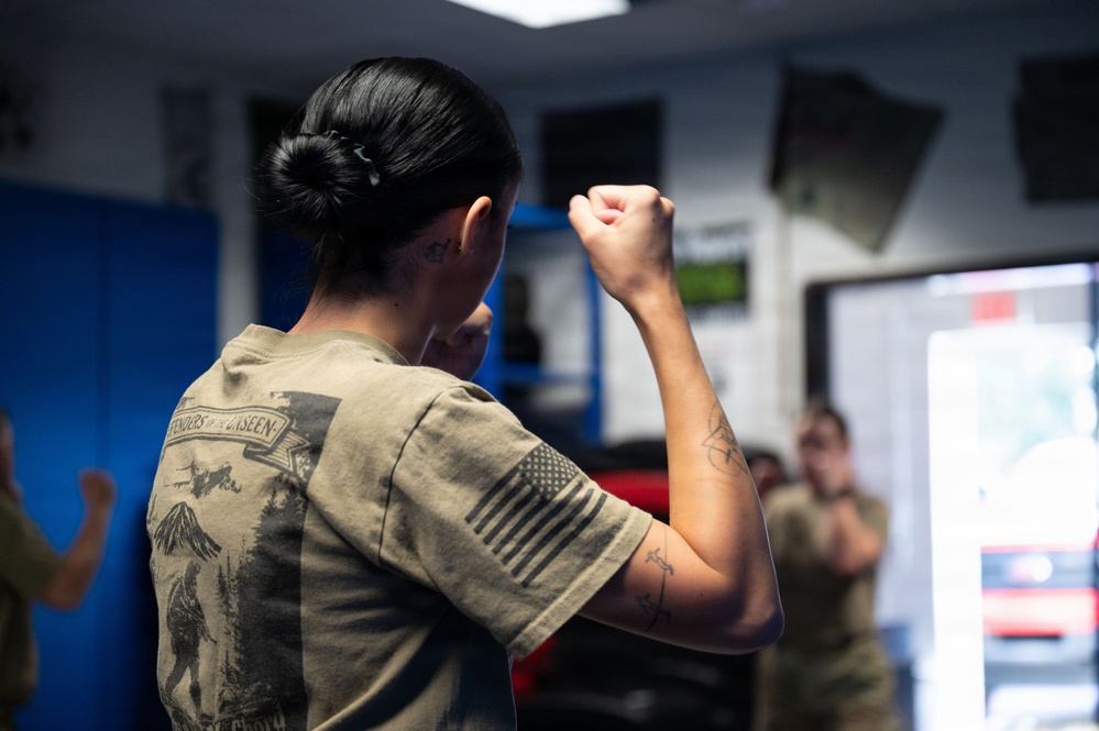 A person wearing an olive/tan military-style t-shirt with an American flag patch on the sleeve is shown from behind with a raised fist in a fighting stance. The setting appears to be an indoor training facility or gym where a group of similarly dressed individuals are practicing combat or self-defense techniques. The foreground subject has visible tattoos on their forearm and neck.