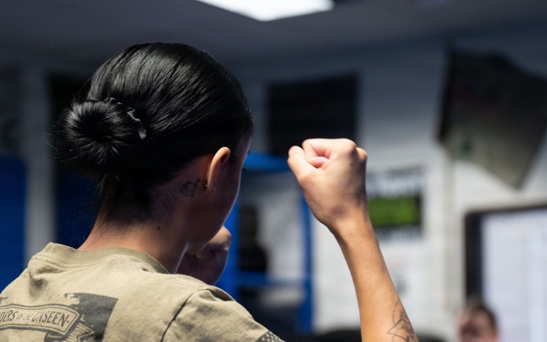 A person wearing an olive/tan military-style t-shirt with an American flag patch on the sleeve is shown from behind with a raised fist in a fighting stance. The setting appears to be an indoor training facility or gym where a group of similarly dressed individuals are practicing combat or self-defense techniques. The foreground subject has visible tattoos on their forearm and neck.