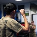 A person wearing an olive/tan military-style t-shirt with an American flag patch on the sleeve is shown from behind with a raised fist in a fighting stance. The setting appears to be an indoor training facility or gym where a group of similarly dressed individuals are practicing combat or self-defense techniques. The foreground subject has visible tattoos on their forearm and neck.
