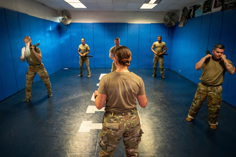 A military or law enforcement training session is taking place in a padded indoor training room. A female instructor, seen from behind, faces five trainees who are holding batons or training sticks in defensive stances. The room features blue padded walls, ceiling fans, and marked floor positions.
