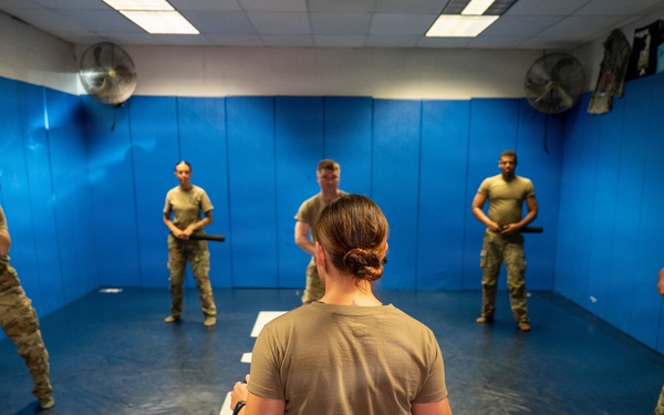 A military or law enforcement training session is taking place in a padded indoor training room. A female instructor, seen from behind, faces five trainees who are holding batons or training sticks in defensive stances. The room features blue padded walls, ceiling fans, and marked floor positions.