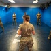 A military or law enforcement training session is taking place in a padded indoor training room. A female instructor, seen from behind, faces five trainees who are holding batons or training sticks in defensive stances. The room features blue padded walls, ceiling fans, and marked floor positions.