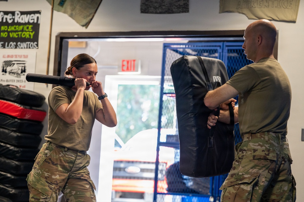 Two military personnel in MultiCam/OCP uniforms are engaged in combatives or hand-to-hand combat training inside what appears to be a martial arts or training facility. A female soldier is in a fighting stance with fists raised, while a male soldier holds a large padded striking shield. The environment includes motivational posters, stacked tires, and training equipment typical of a combat sports gym.