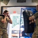 Two military personnel in MultiCam/OCP uniforms are engaged in combatives or hand-to-hand combat training inside what appears to be a martial arts or training facility. A female soldier is in a fighting stance with fists raised, while a male soldier holds a large padded striking shield. The environment includes motivational posters, stacked tires, and training equipment typical of a combat sports gym.