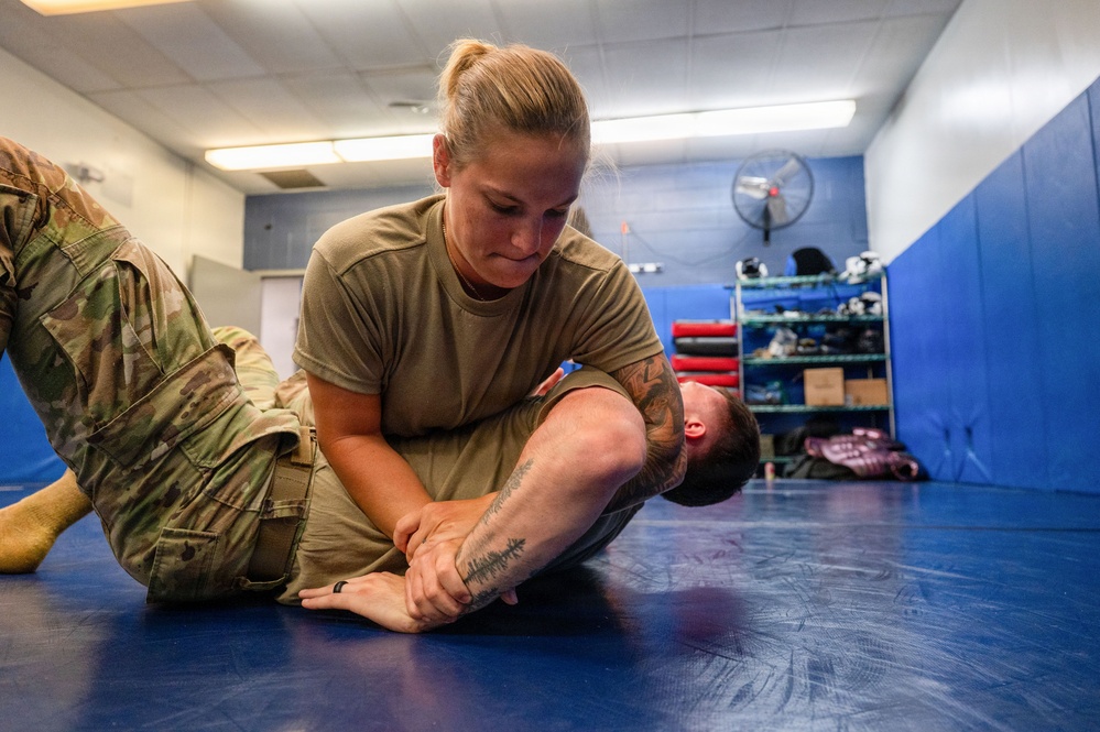 Two military personnel in camouflage uniforms are practicing ground combat or combatives training on a blue mat in an indoor facility. A female soldier is applying a grappling hold or submission technique on a male soldier who is on the ground. The setting appears to be a dedicated combatives or martial arts training room.