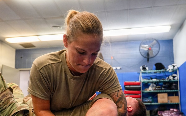Two military personnel in camouflage uniforms are practicing ground combat or combatives training on a blue mat in an indoor facility. A female soldier is applying a grappling hold or submission technique on a male soldier who is on the ground. The setting appears to be a dedicated combatives or martial arts training room.