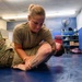 Two military personnel in camouflage uniforms are practicing ground combat or combatives training on a blue mat in an indoor facility. A female soldier is applying a grappling hold or submission technique on a male soldier who is on the ground. The setting appears to be a dedicated combatives or martial arts training room.