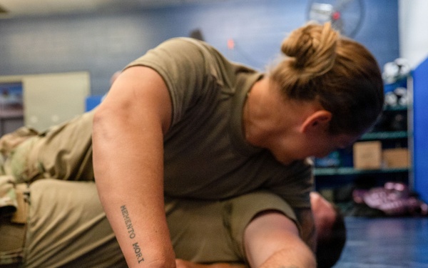 Two military personnel in olive/camouflage uniforms are engaged in a ground combat or combatives training exercise on a blue mat. One individual is applying a ground control technique, pinning the other person's arm while in a dominant top position. The setting appears to be an indoor military training facility or gym.