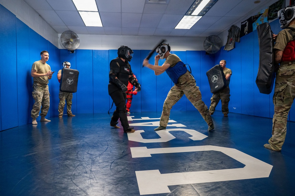 Military or law enforcement personnel are conducting combatives/defensive tactics training in a padded training facility. One participant in full protective gear (black suit, helmet) is engaged in a baton/stick drill against another participant wearing a blue vest and protective headgear. Observers in military camouflage uniforms watch from the perimeter, some holding training shields.