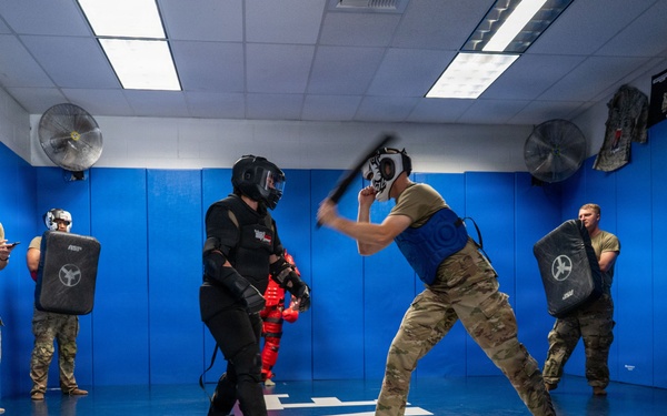 Military or law enforcement personnel are conducting combatives/defensive tactics training in a padded training facility. One participant in full protective gear (black suit, helmet) is engaged in a baton/stick drill against another participant wearing a blue vest and protective headgear. Observers in military camouflage uniforms watch from the perimeter, some holding training shields.