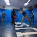 Military or law enforcement personnel are conducting combatives/defensive tactics training in a padded training facility. One participant in full protective gear (black suit, helmet) is engaged in a baton/stick drill against another participant wearing a blue vest and protective headgear. Observers in military camouflage uniforms watch from the perimeter, some holding training shields.