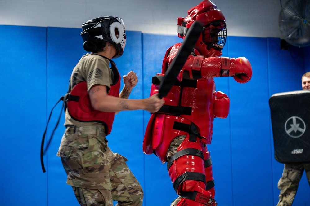 A military combatives training session is taking place in an indoor padded facility. A person in military camouflage (MultiCam/OCP uniform) wearing a protective helmet and red chest guard is practicing strikes against an instructor wearing full red padded protective suit (FIST suit) and holding a padded baton. A third person is visible in the background holding a strike shield.