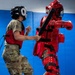 A military combatives training session is taking place in an indoor padded facility. A person in military camouflage (MultiCam/OCP uniform) wearing a protective helmet and red chest guard is practicing strikes against an instructor wearing full red padded protective suit (FIST suit) and holding a padded baton. A third person is visible in the background holding a strike shield.