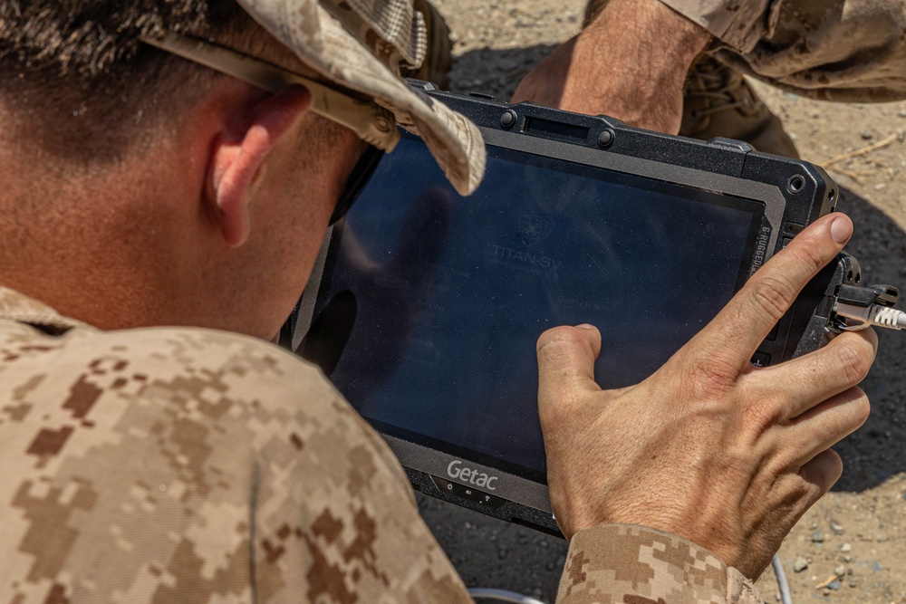 A military personnel wearing desert camouflage (MARPAT pattern) uniform and cap is operating a Getac rugged tablet in an outdoor environment. The tablet displays 'TITAN-5V' on its screen and is connected via a cable. The setting appears to be an arid or semi-arid outdoor location.