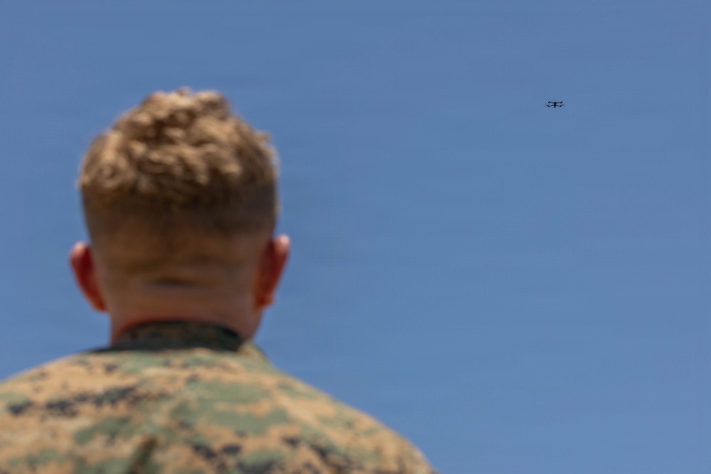 A military personnel member, visible from behind, is observing a small drone or UAV flying in a clear blue sky. The individual is wearing a camouflage uniform consistent with U.S. Marine Corps MARPAT pattern. The drone is visible in the upper right portion of the frame at a significant distance.