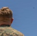 A military personnel member, visible from behind, is observing a small drone or UAV flying in a clear blue sky. The individual is wearing a camouflage uniform consistent with U.S. Marine Corps MARPAT pattern. The drone is visible in the upper right portion of the frame at a significant distance.