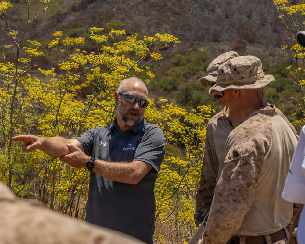 A civilian contractor or advisor wearing a dark polo shirt with 'DZYN' branding is briefing or directing two military personnel in desert camouflage (MARPAT) uniforms and boonie hats in an outdoor, arid environment. The civilian is pointing in a specific direction, suggesting a terrain briefing or site assessment. The setting appears to be a rocky, semi-arid hillside with yellow flowering plants in the background.