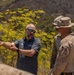 A civilian contractor or advisor wearing a dark polo shirt with 'DZYN' branding is briefing or directing two military personnel in desert camouflage (MARPAT) uniforms and boonie hats in an outdoor, arid environment. The civilian is pointing in a specific direction, suggesting a terrain briefing or site assessment. The setting appears to be a rocky, semi-arid hillside with yellow flowering plants in the background.