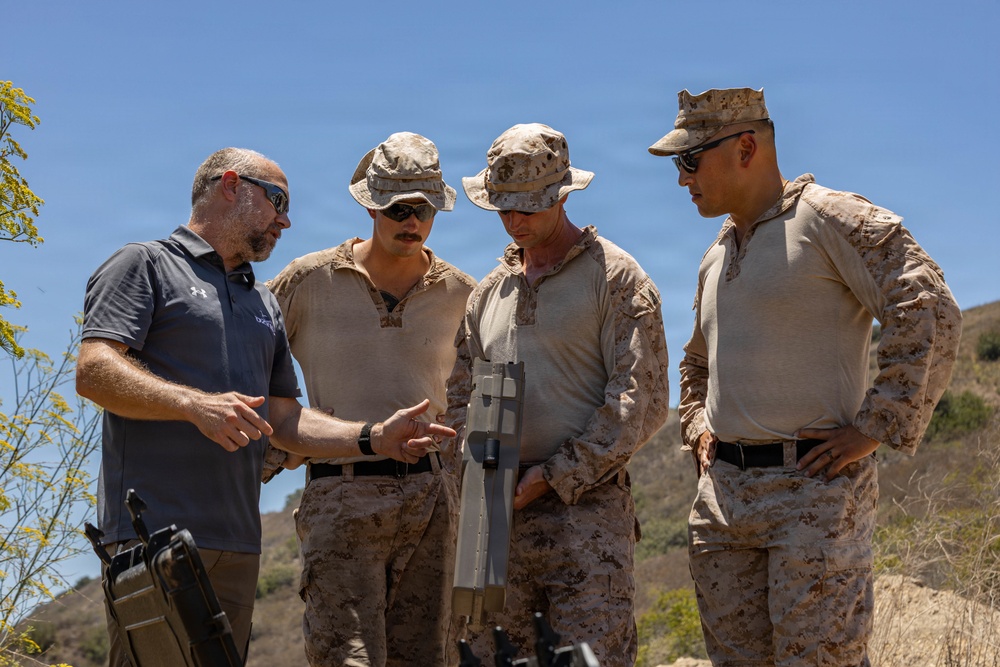 Four individuals, three in desert camouflage military uniforms and one in civilian attire, are gathered outdoors examining a piece of equipment. The civilian, wearing a dark polo shirt with what appears to be a company logo, appears to be briefing or demonstrating the equipment to the military personnel. The setting appears to be a training or demonstration area in a dry, hilly terrain under clear blue skies.