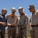 Four individuals, three in desert camouflage military uniforms and one in civilian attire, are gathered outdoors examining a piece of equipment. The civilian, wearing a dark polo shirt with what appears to be a company logo, appears to be briefing or demonstrating the equipment to the military personnel. The setting appears to be a training or demonstration area in a dry, hilly terrain under clear blue skies.