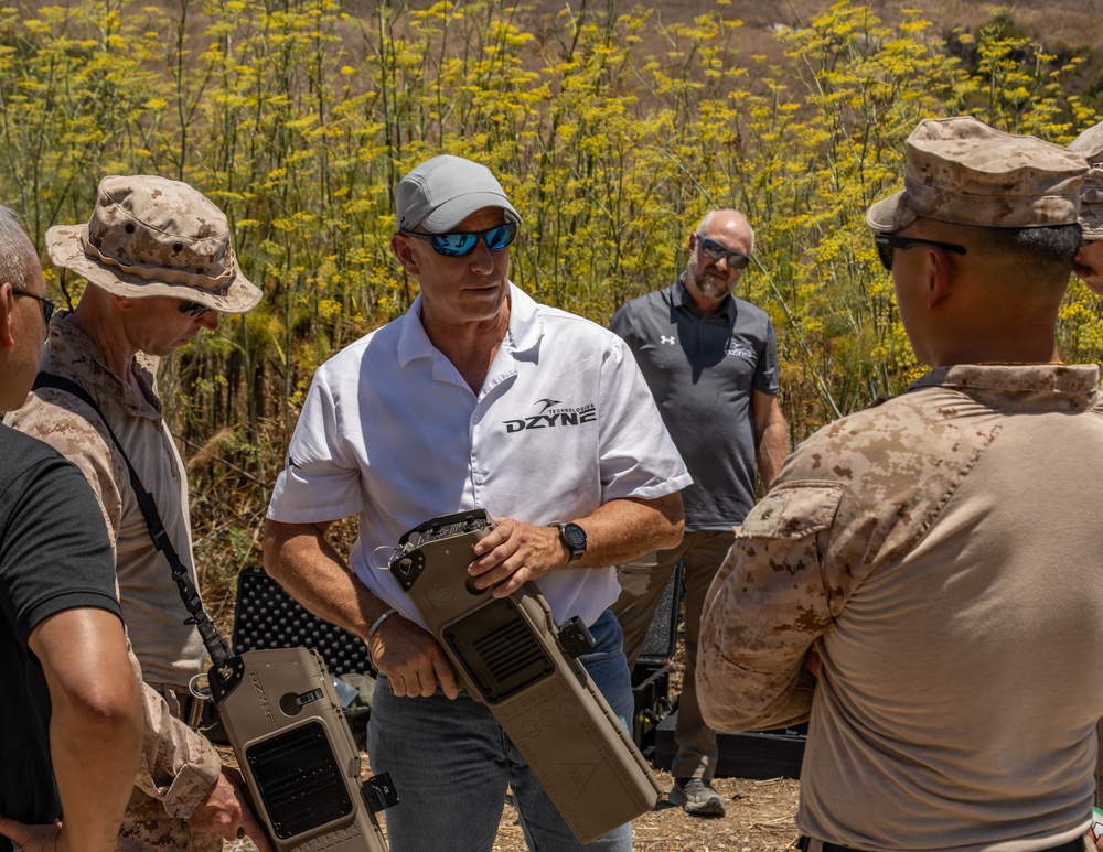 A representative from DZYNE Technologies is demonstrating equipment to what appear to be U.S. Marines in a field environment. The civilian in a white DZYNE polo shirt is holding a tan-colored tactical case or equipment unit while briefing military personnel in desert camouflage uniforms. A second DZYNE representative in a gray shirt observes in the background.