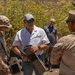 A representative from DZYNE Technologies is demonstrating equipment to what appear to be U.S. Marines in a field environment. The civilian in a white DZYNE polo shirt is holding a tan-colored tactical case or equipment unit while briefing military personnel in desert camouflage uniforms. A second DZYNE representative in a gray shirt observes in the background.